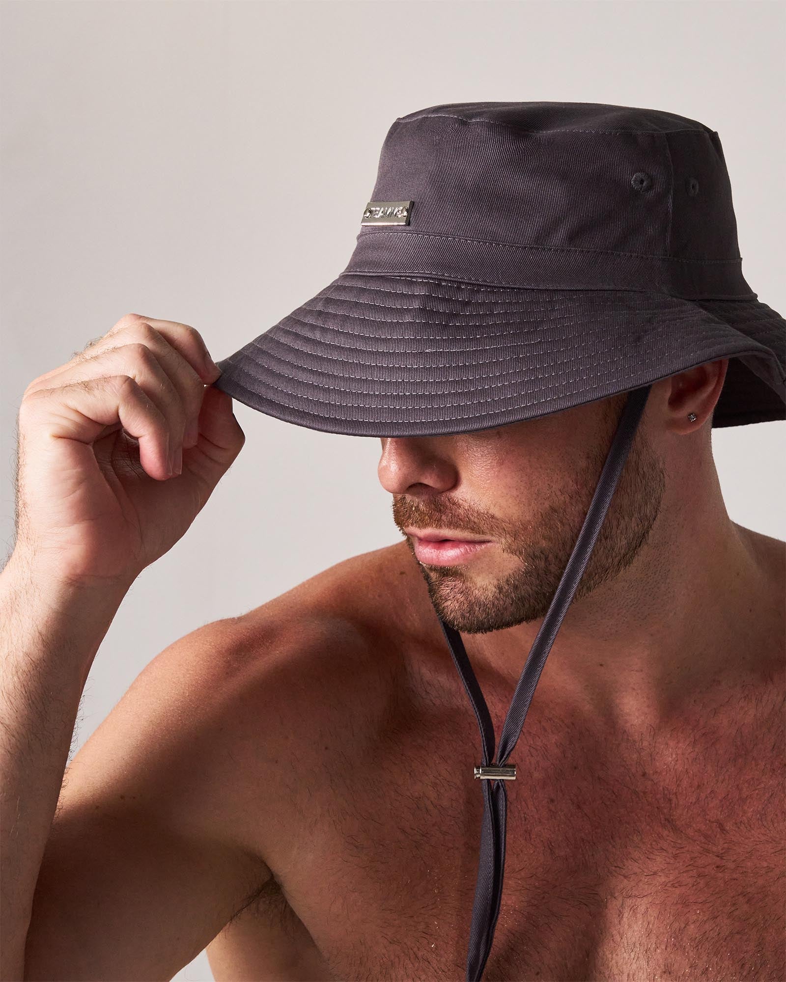 Man wearing a dark gray bucket hat with a strap on a plain background
